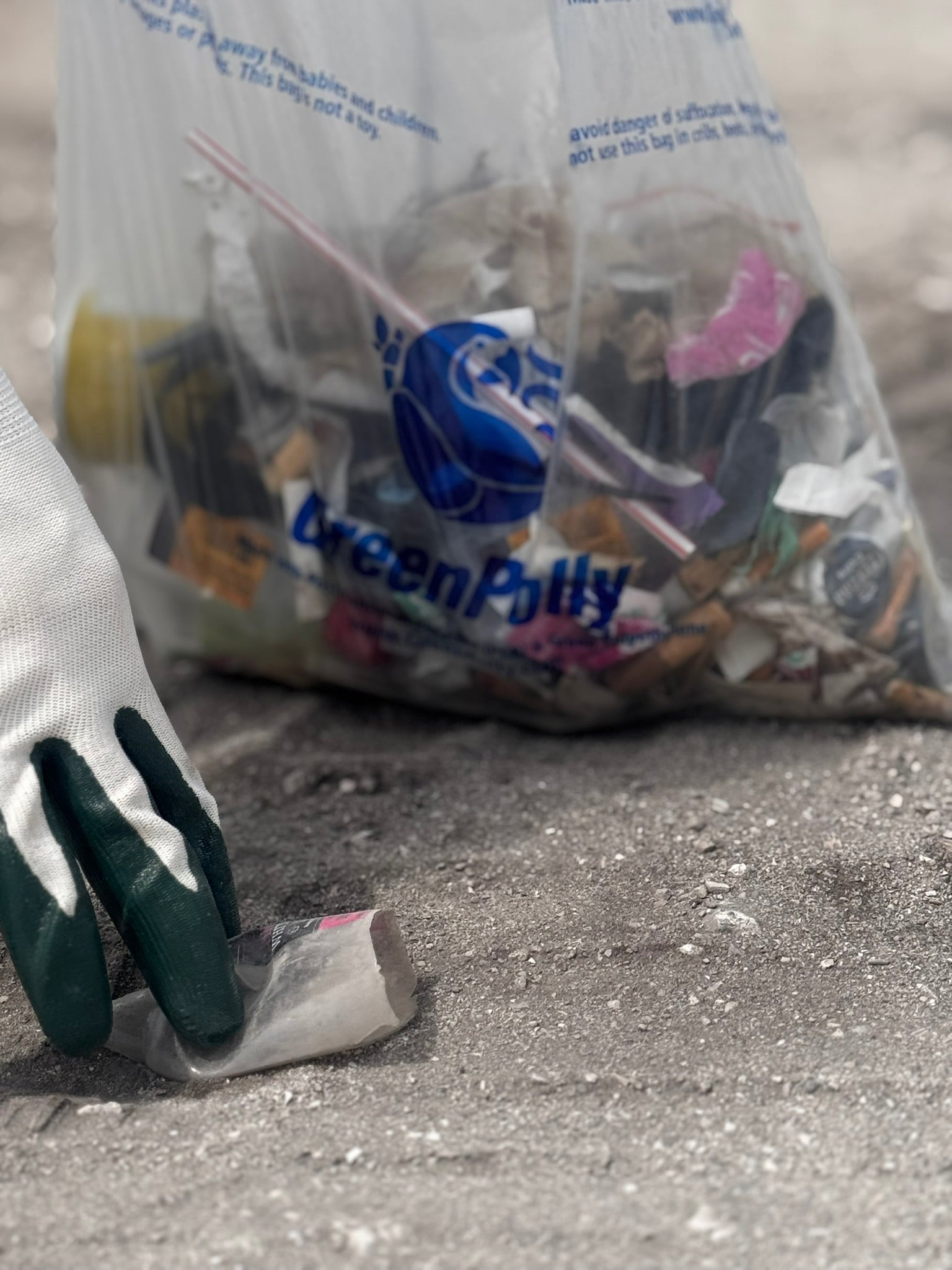 A gloved hand places a small plastic item into a large clear recycling bag labeled GreenPoly, set on a gray concrete surface with the bag blurred in the background.